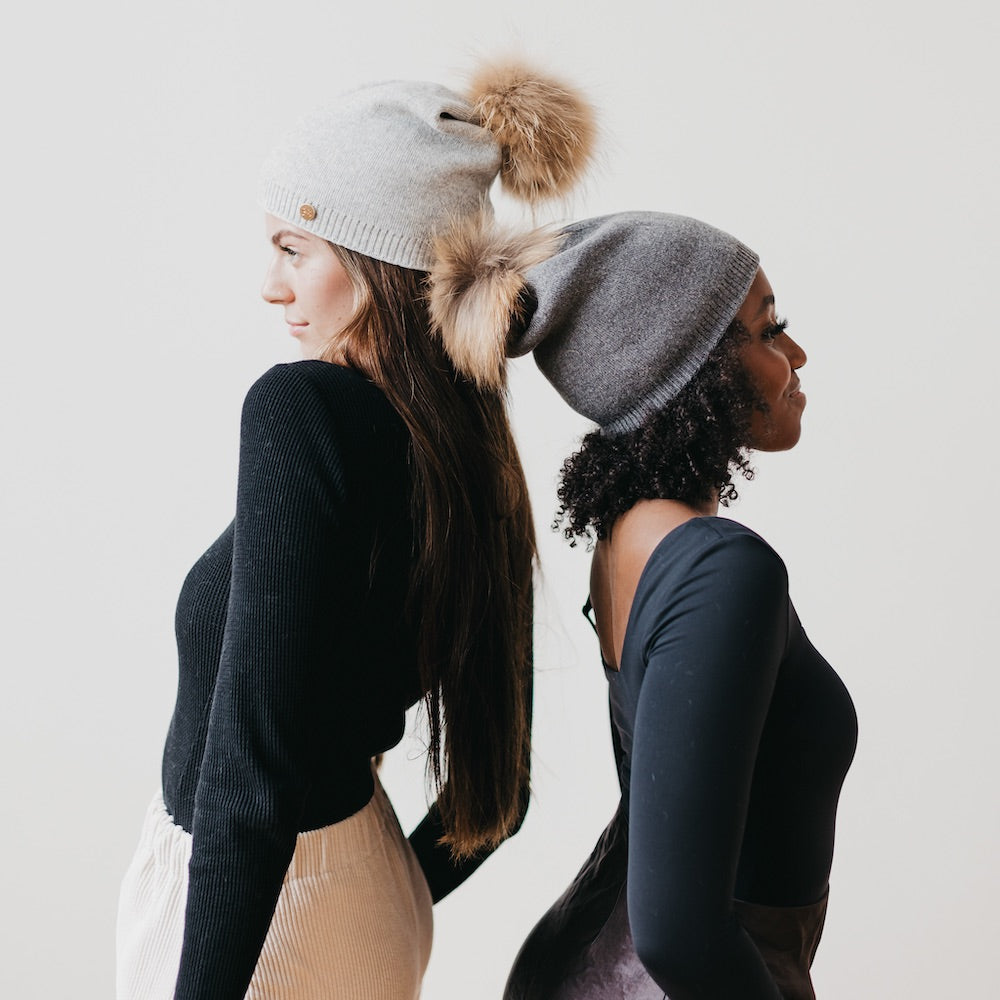 Two women wearing gray knit hats with pom-poms against a plain background