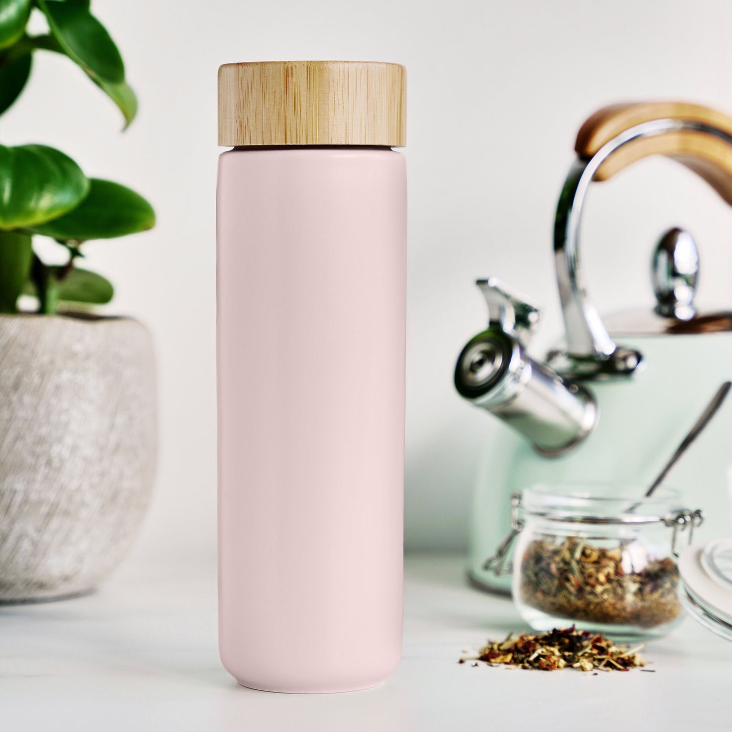 Pink Tea infuser with wooden lid on a white surface with a plant and teapot in the background