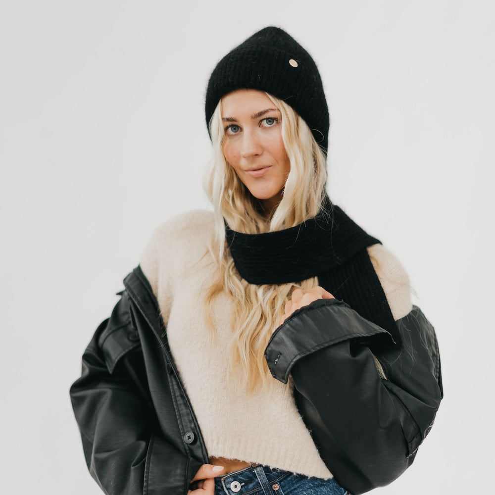 Woman wearing a black knit beanie, black scarf, and black leather jacket on a light gray background