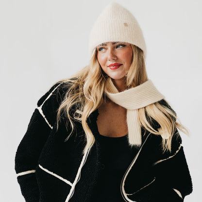 Woman wearing a black coat with white trim, cream beanie, and scarf on a light gray background
