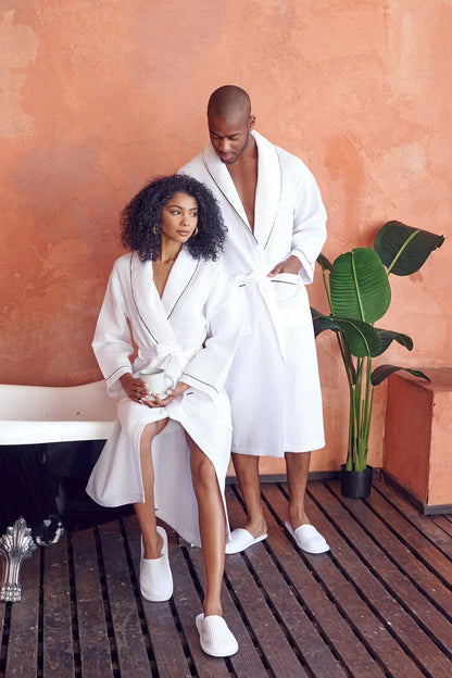 Two people in white waffle robes standing in a bathroom with a bathtub and plant.