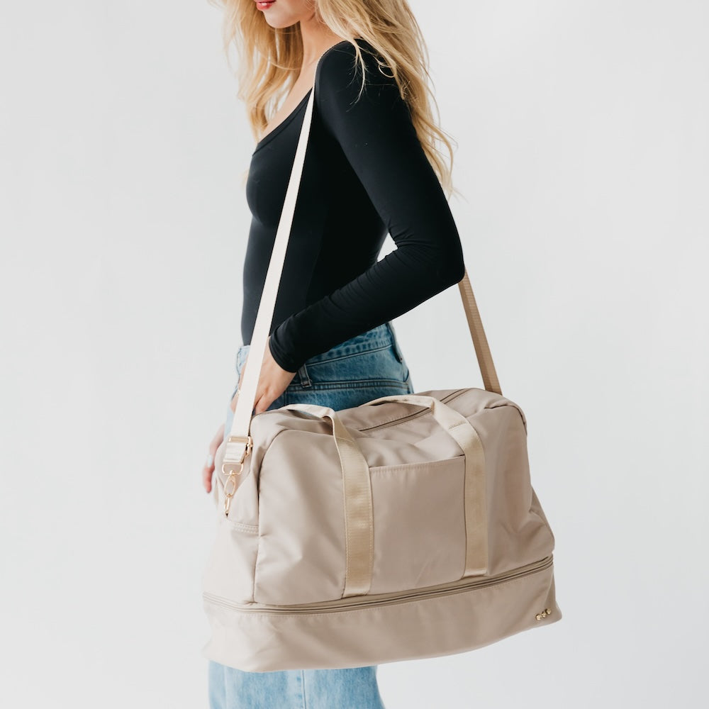 Woman carrying a beige duffel bag on a plain background