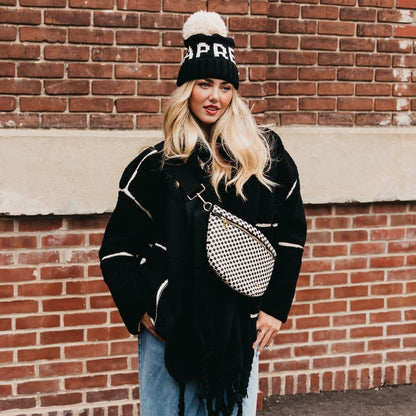 Woman wearing a black coat, jeans, and a beanie with a checkered bag against a brick wall.