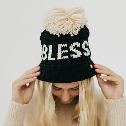 Black knit beanie with 'BLESS' text and white pom-pom held by a person against a light background