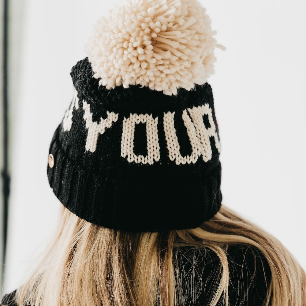 Black knit beanie with a large pom-pom worn by a person with blonde hair.