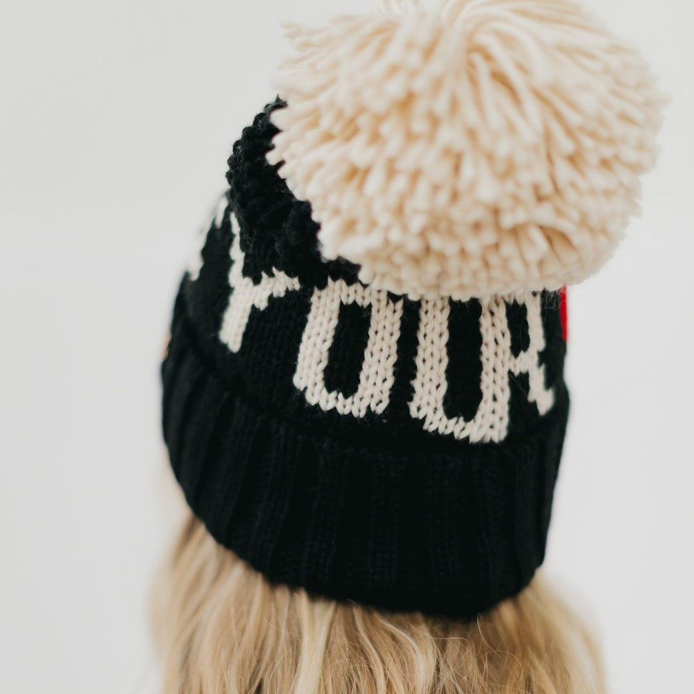 Black knit beanie with a beige pom-pom on a person wearing it.
