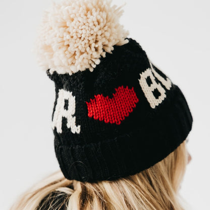 Black knit beanie with a red heart and white text, worn by a person with blonde hair.