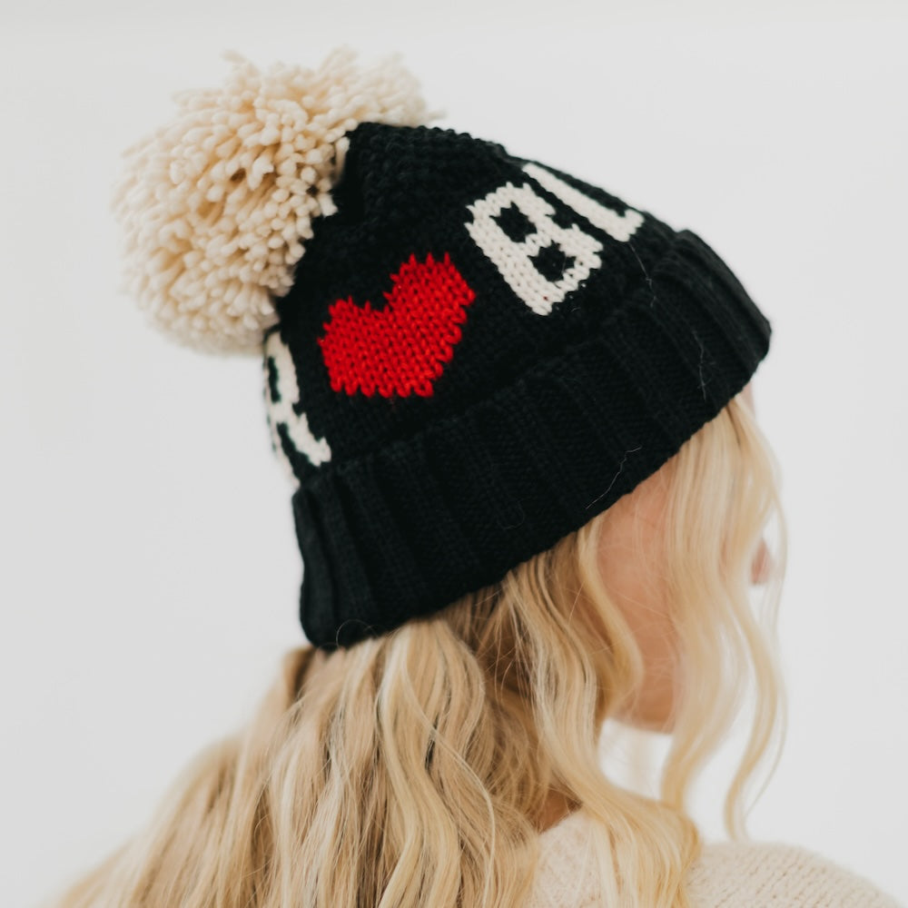 Black knit beanie with red heart and white text, worn by a person with blonde hair.