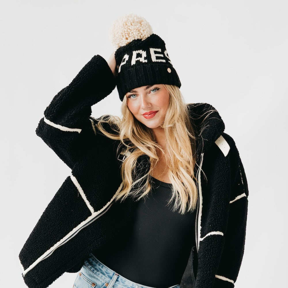Woman wearing a black beanie with pom-pom and a matching black cardigan on a light gray background