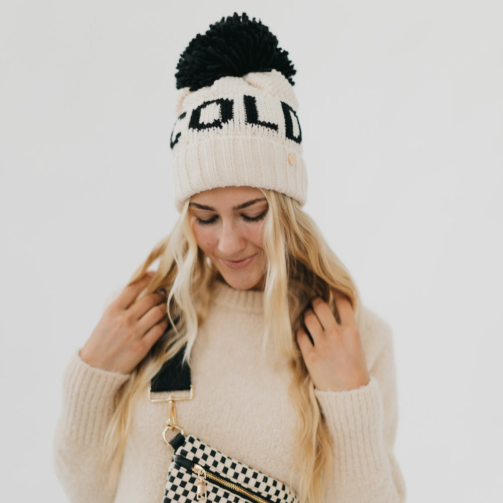 Person wearing a white knit beanie with black pom-pom and 'OLD' text, holding a checkered bag.