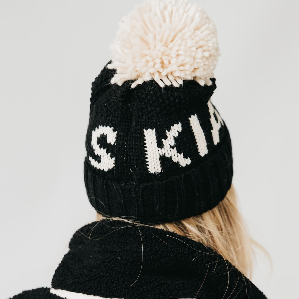 Black knit beanie with white text and pom-pom on a plain background