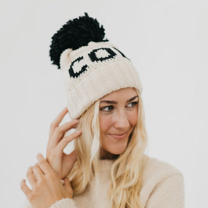 Woman wearing a cream beanie with black pom-pom and 'CO' text on a white background