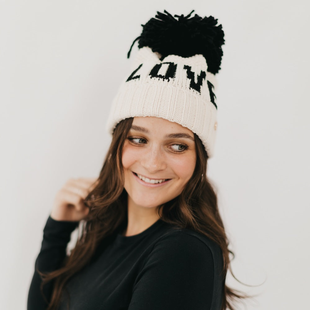Person wearing a white beanie with black pom-poms and 'LOVE' text on a plain background