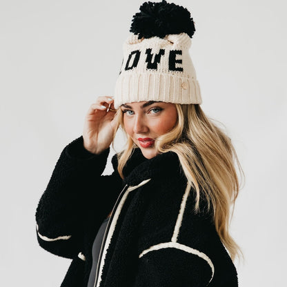 Woman wearing a black and white coat and a cream beanie with 'LOVE' and a pom-pom on a light gray background