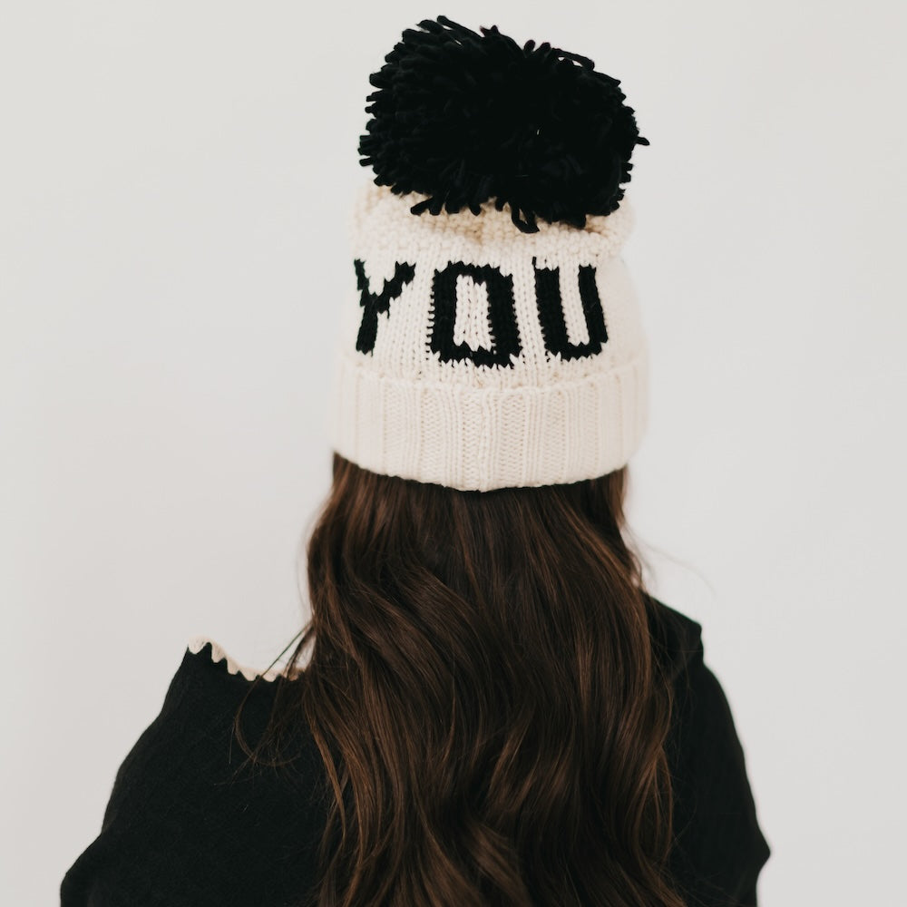 Person wearing a white knit beanie with black text and pom-pom on a plain background