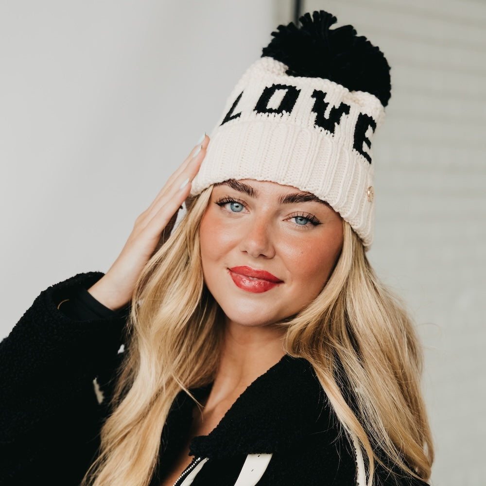 Woman wearing a white knit beanie with black text and pom-poms, against a plain background.