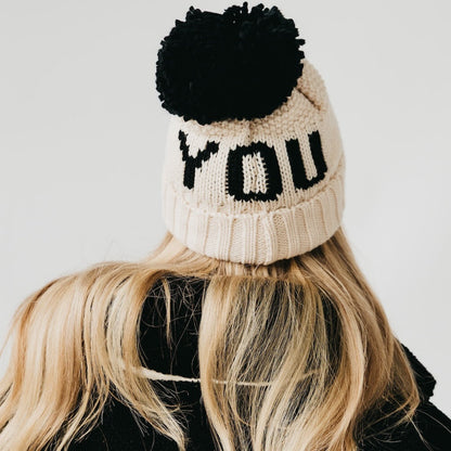 Beige knit beanie with black pom-pom and 'YOU' text on a plain background