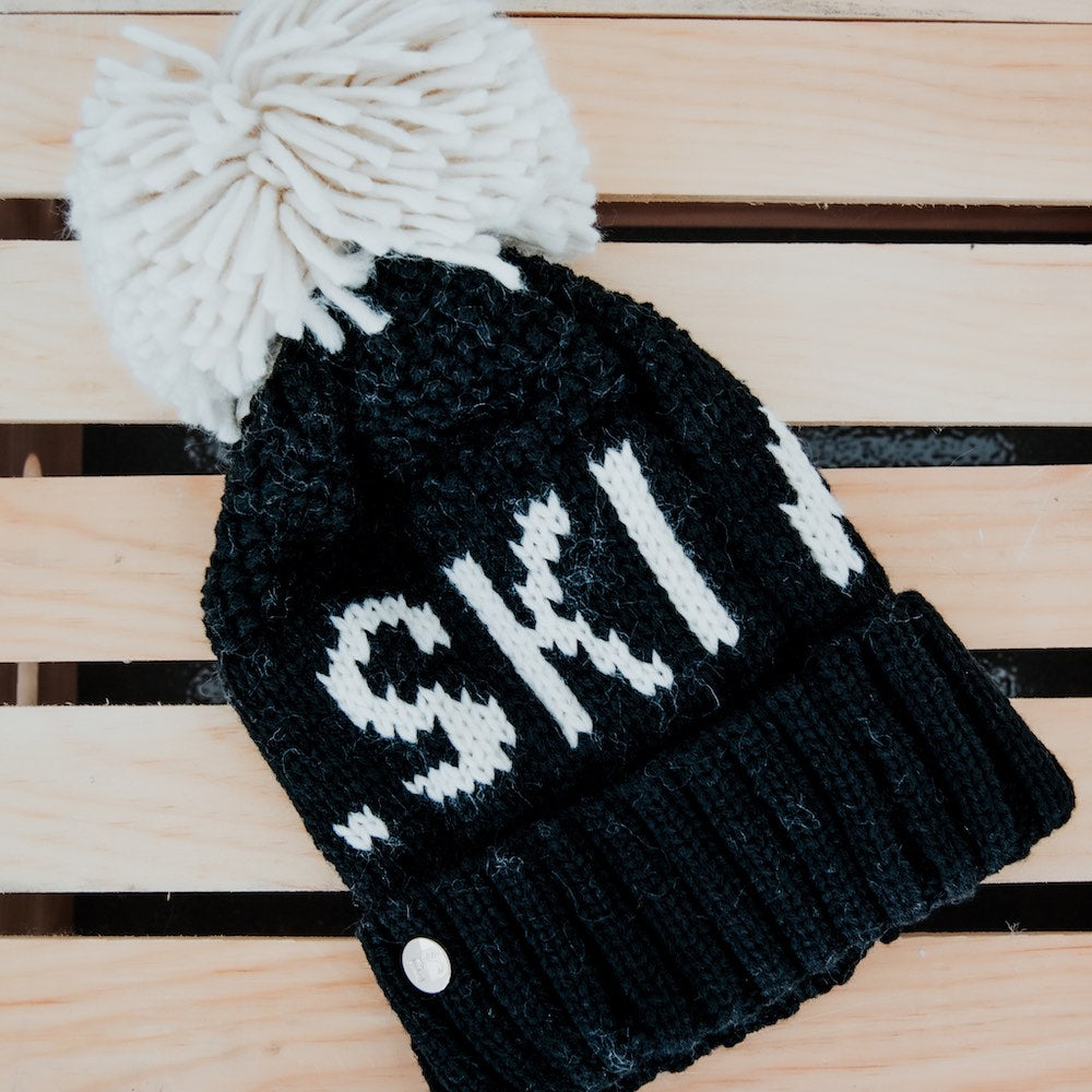 Black knit beanie with white pom-pom and 'Ski!' text on a wooden surface