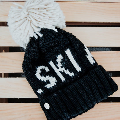 Black knit beanie with white pom-pom and 'Ski!' text on a wooden surface