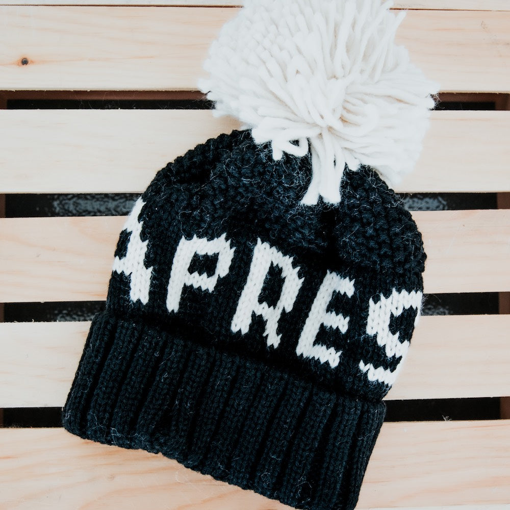 Black knit beanie with white text and pom-pom on a wooden surface