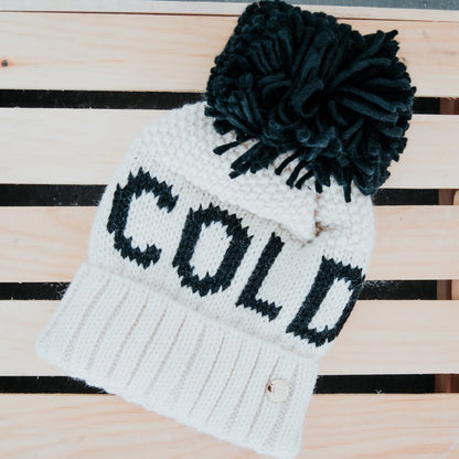 White knit beanie with black text 'COLD' and pom-pom on a striped wooden background