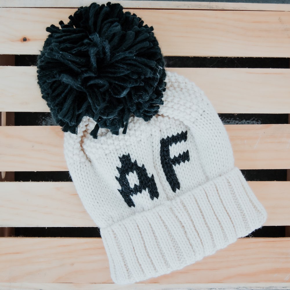 White knit beanie with black pom-pom and 'AF' text on a wooden surface