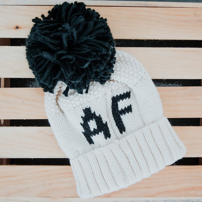 White knit beanie with black pom-pom and 'AF' text on a wooden surface