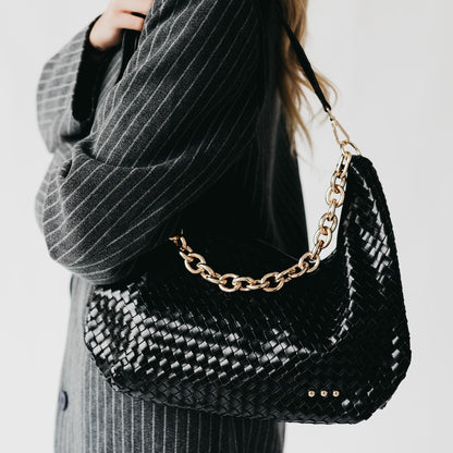 Black woven handbag with gold chain strap held by a person wearing a gray pinstripe outfit.