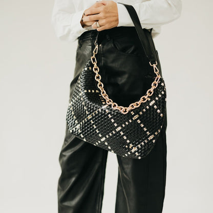 Black woven handbag with gold chain detail held by a person wearing a white shirt and black pants.