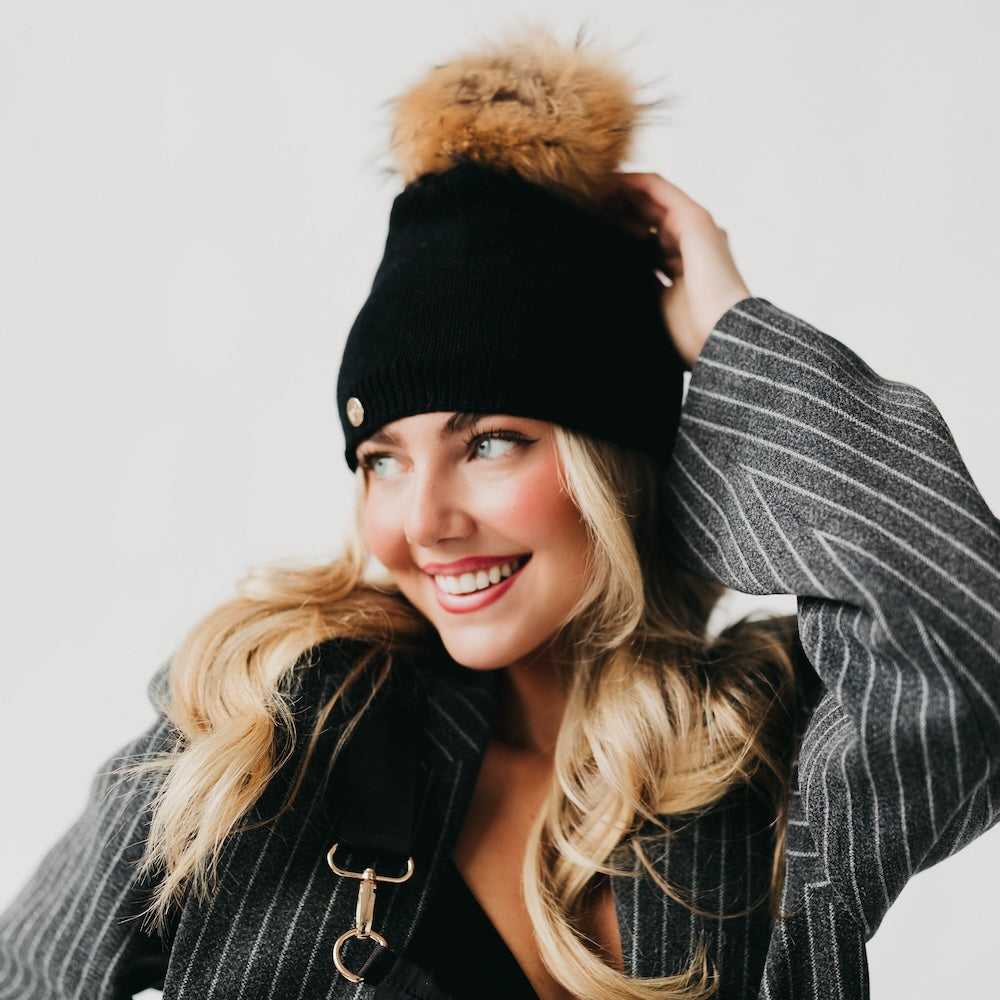 Woman wearing a black beanie with a fur pom-pom and gray pinstripe coat on a light background