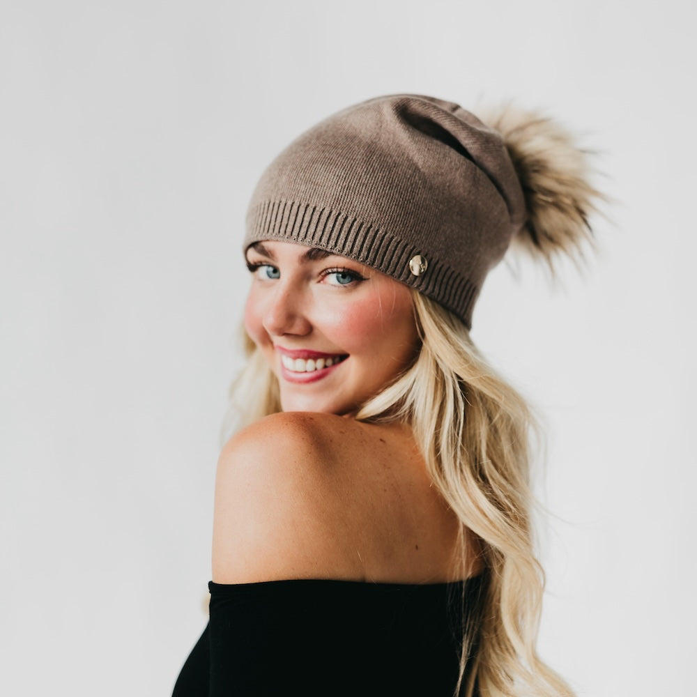 Woman wearing a brown knit beanie with a fluffy pom-pom on a plain background
