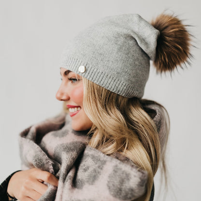 Woman wearing a gray knit beanie with a brown pom-pom and a gray coat against a light background