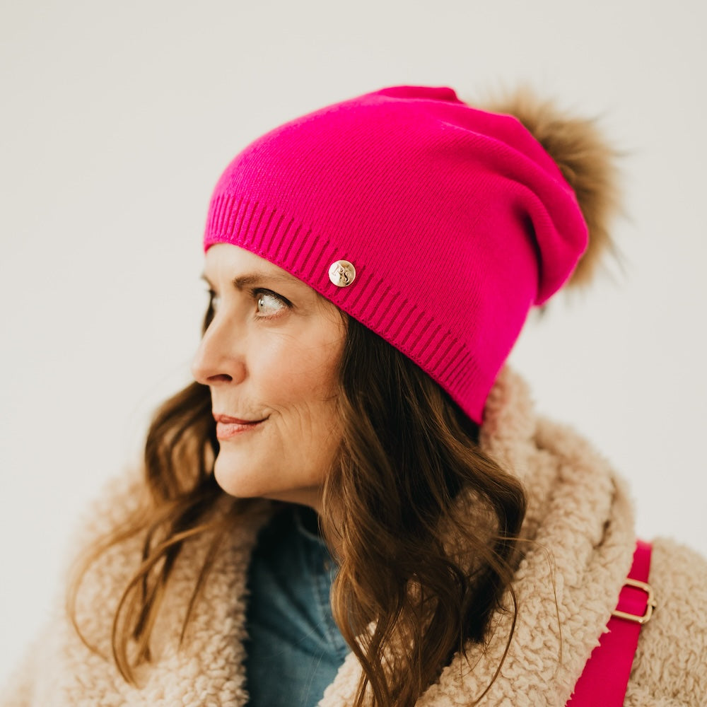 Woman wearing a bright pink beanie with a fluffy pom-pom on a plain background