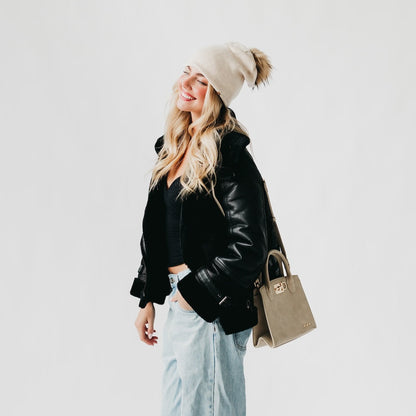 Woman wearing a black leather jacket, light blue jeans, and a beige beanie with a handbag on a white background