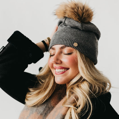 Woman wearing a gray knit beanie with a pom-pom, smiling against a white background