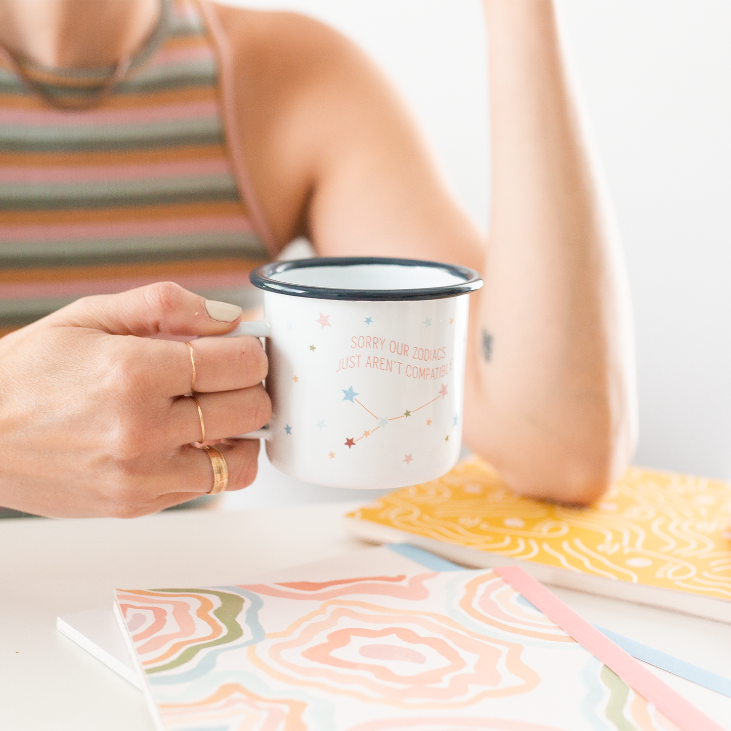 Person holding a mug with text and star designs, sitting at a table with a colorful notebook.