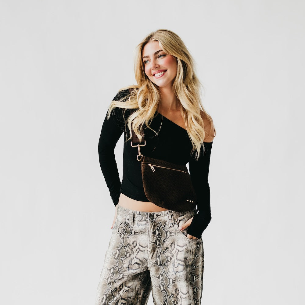 Woman wearing a black long-sleeve top and patterned pants with a brown bag on a light gray background