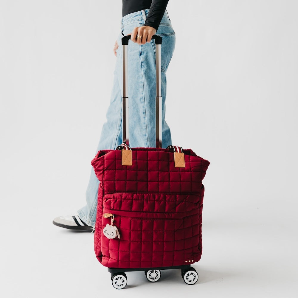 Red quilted suitcase with wheels being held by a person wearing jeans and a black top on a light gray background.