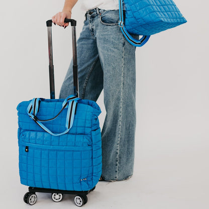 Blue quilted rolling bag held by a person wearing jeans on a light gray background