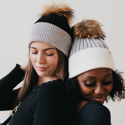 Two women wearing beanies with pom-poms against a plain background