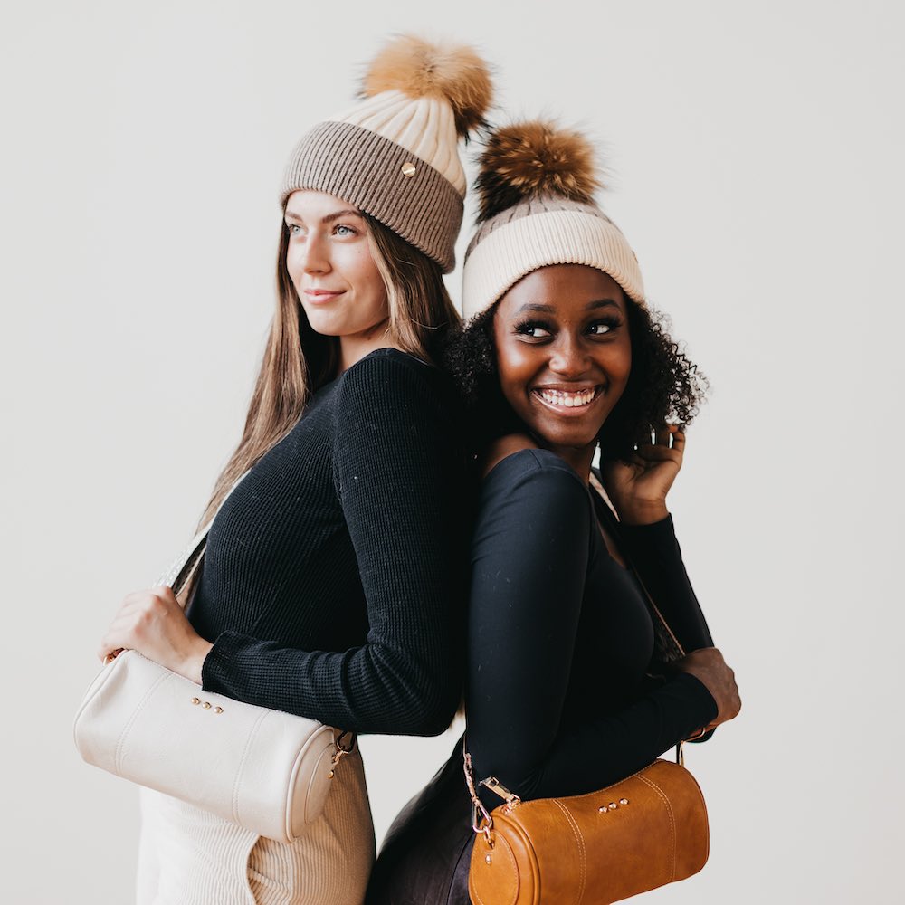Two women wearing matching beanies with pom-poms, one holding a beige handbag and the other a brown handbag, against a plain background.
