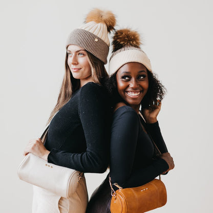 Two women wearing matching beanies with pom-poms, one holding a beige handbag and the other a brown handbag, against a plain background.