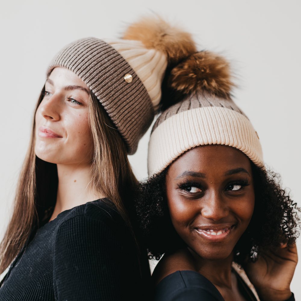 Two women wearing knit hats with pom-poms against a plain background