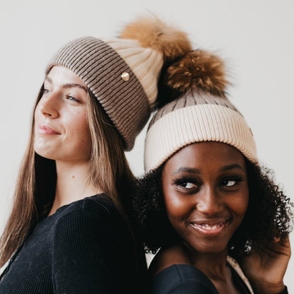Two women wearing knit hats with pom-poms against a plain background