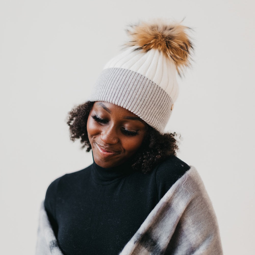Person wearing a white and gray beanie with a brown pom-pom, black turtleneck, and plaid scarf against a plain background.