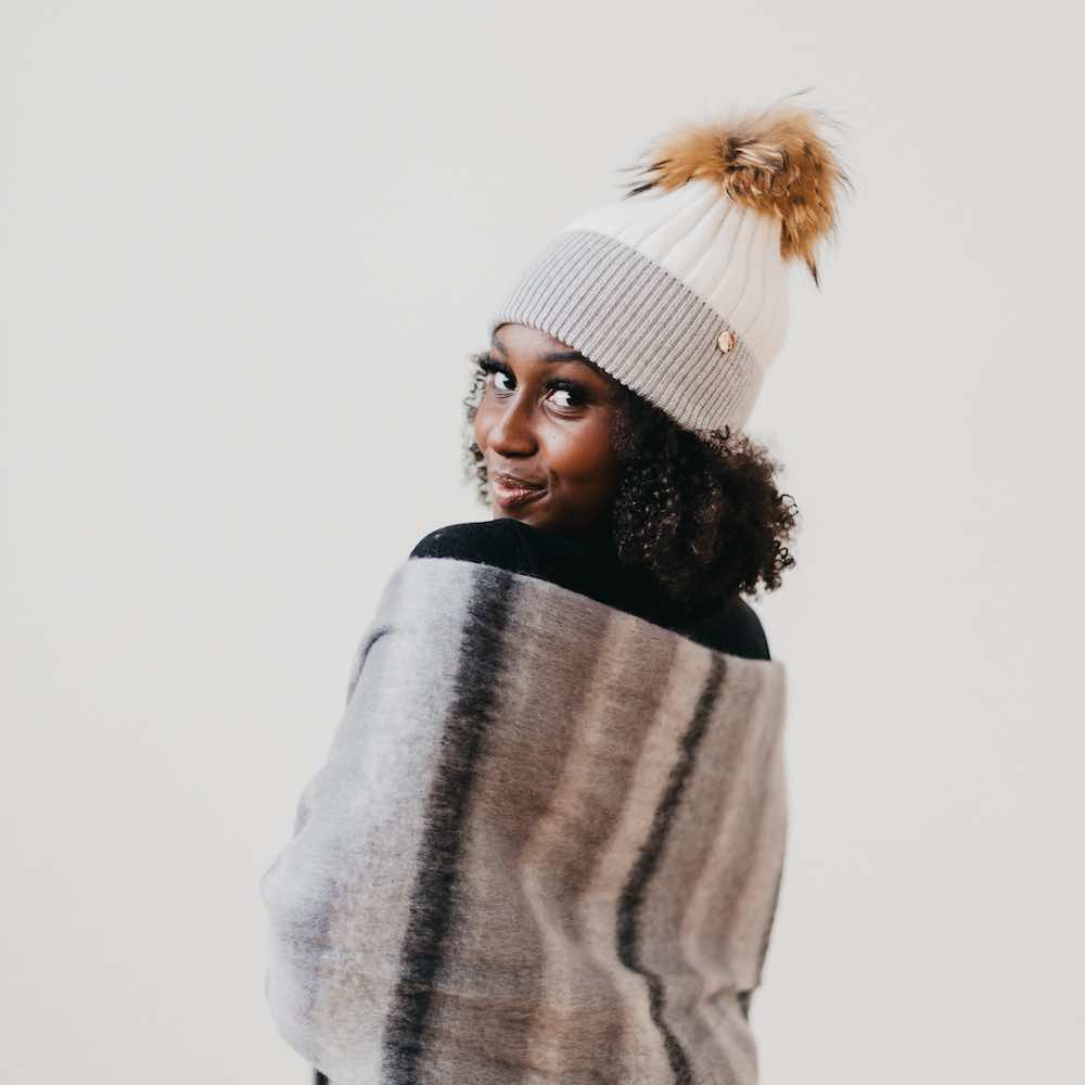 Person wearing a gray knit beanie with a pom-pom and a striped sweater against a white background
