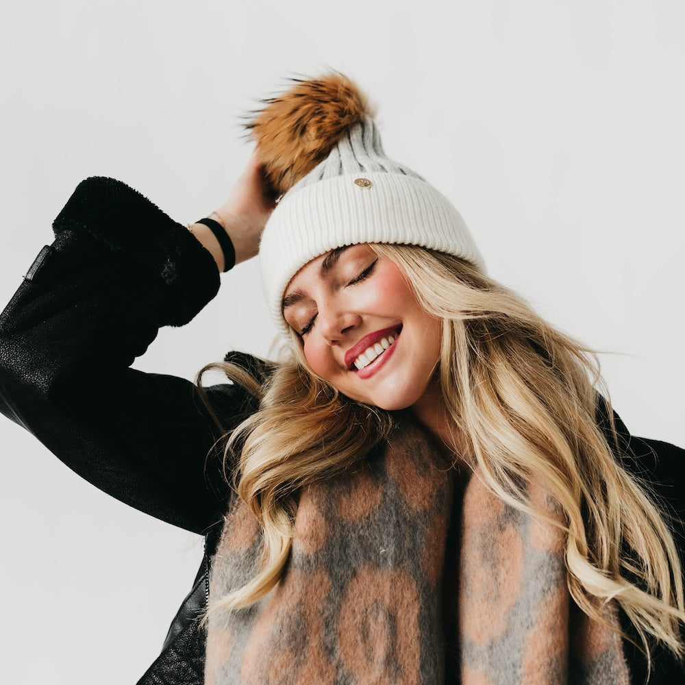 Woman wearing a white beanie with a brown pom-pom and a patterned scarf, smiling against a light background.