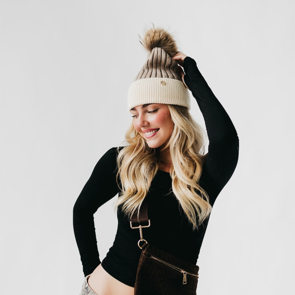 Woman wearing a black long-sleeve top and brown suspenders with a white and beige beanie on a light gray background