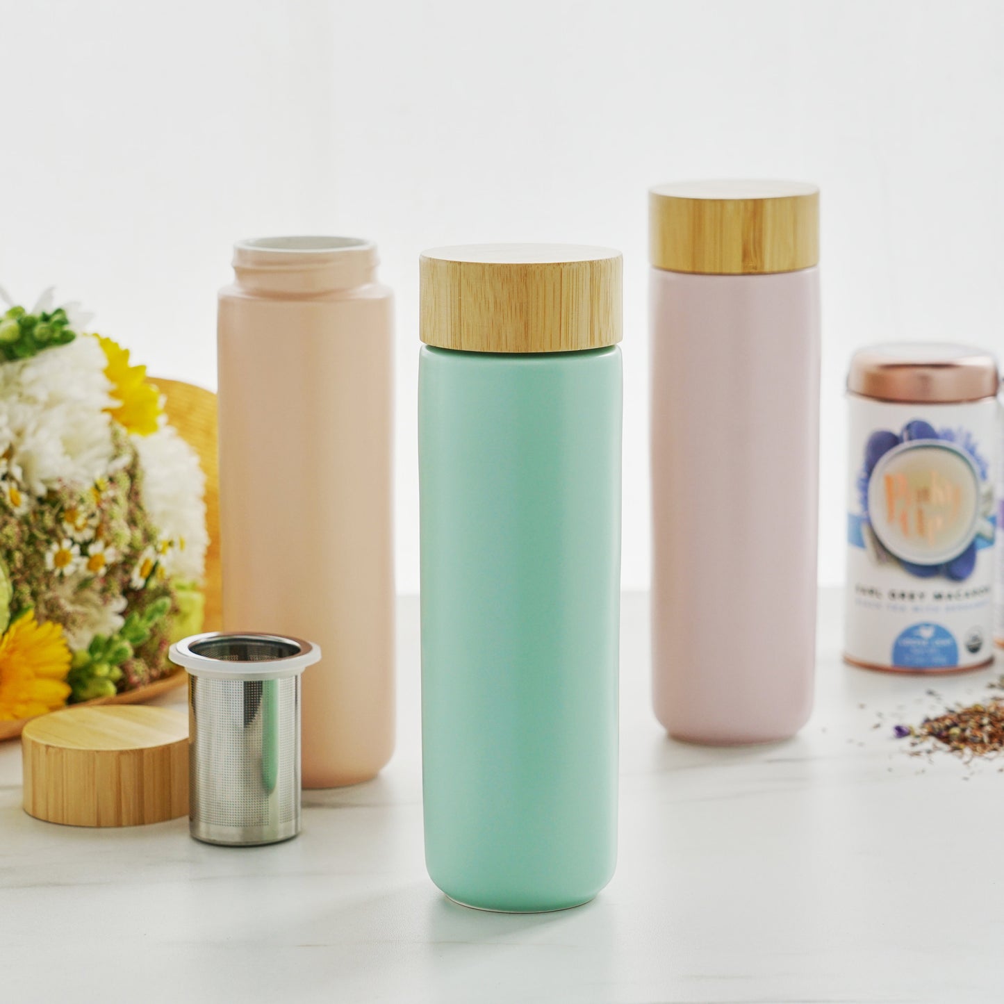 Three colorful to go Tea infuser bottles with wooden lids on a white surface with flowers and a jar in the background.
