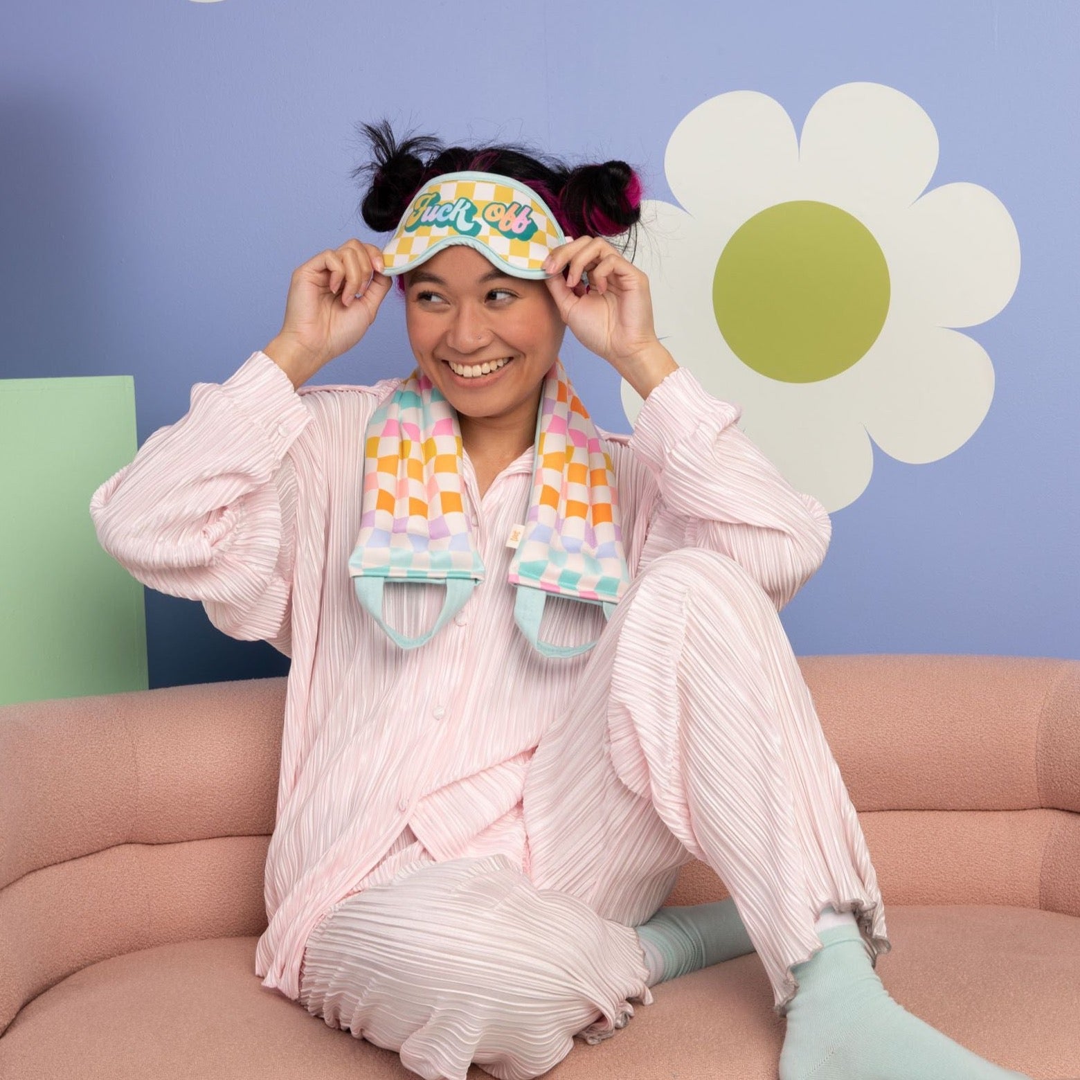 Person wearing a colorful eye mask and scarf, sitting on a couch with a playful background.